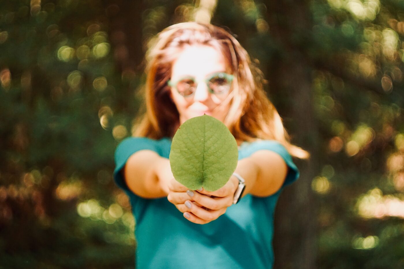 A person holding a large green leaf towards the camera, partially obscuring their face in front of a forest.