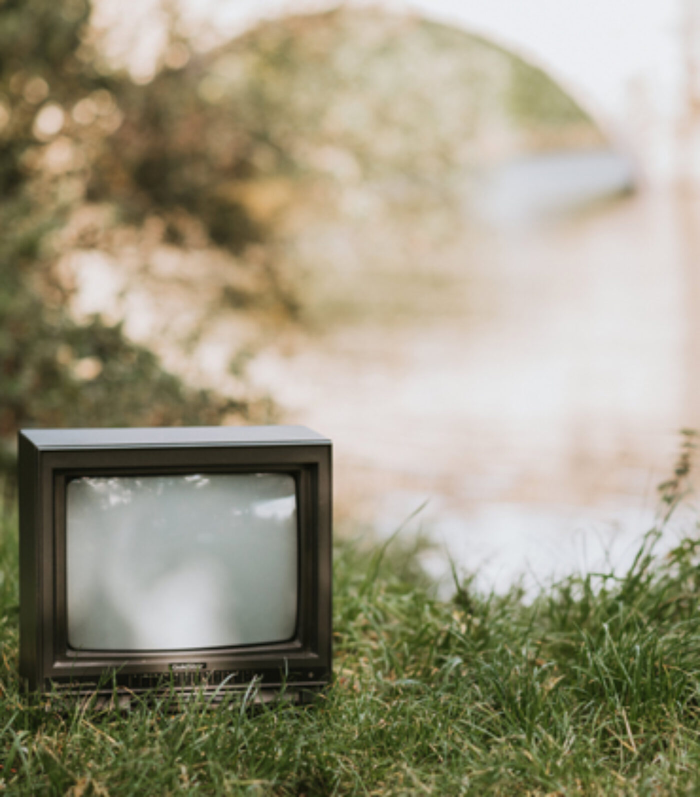 An old, small television set sits on the grass near a blurred, sunlit background of trees and a bridge.