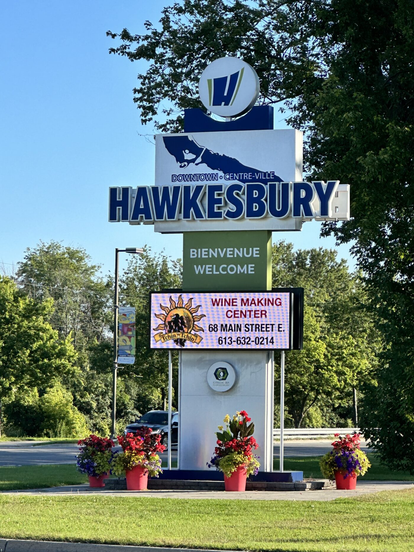 Photo of the welcome sign in downtown Hawkesbury accompanied by an outdoor advertising screen, surrounded by flower pots