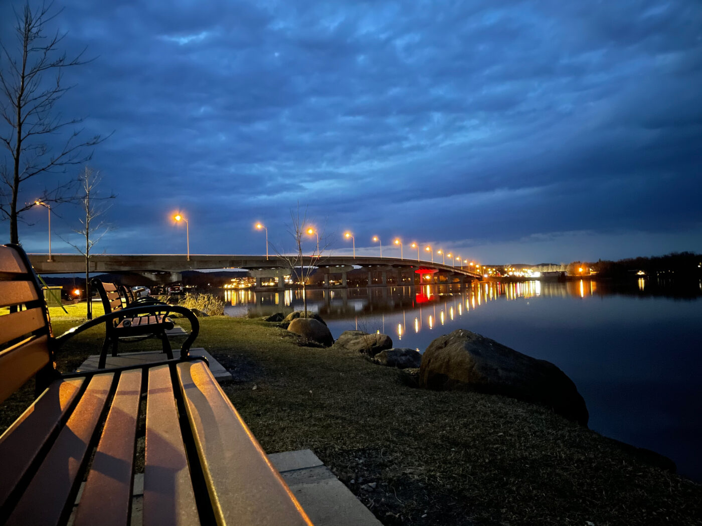 Photo of public benches on the riverbank at dusk, with a bridge in the background