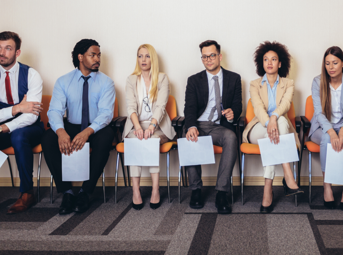 Photo of colleagues seated on orange chairs in a line, each holding a document.